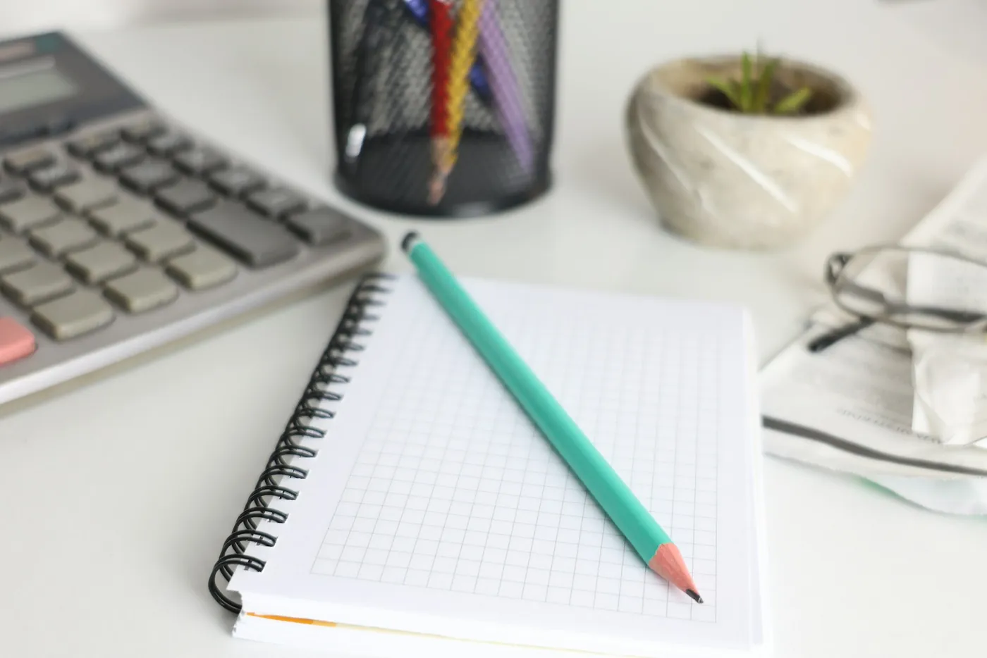 A desk calculator, a grid notebook with a green pencil, and a small potted plant on a white desk